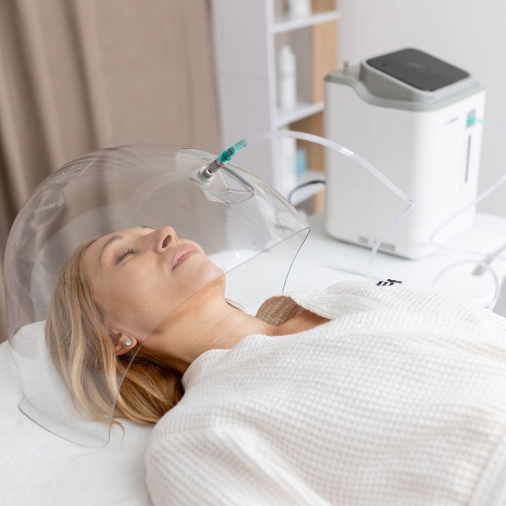 Woman relaxing under a clear oxygen dome attached to a skincare oxygen machine during an oxygen infusion facial session in a treatment room.
