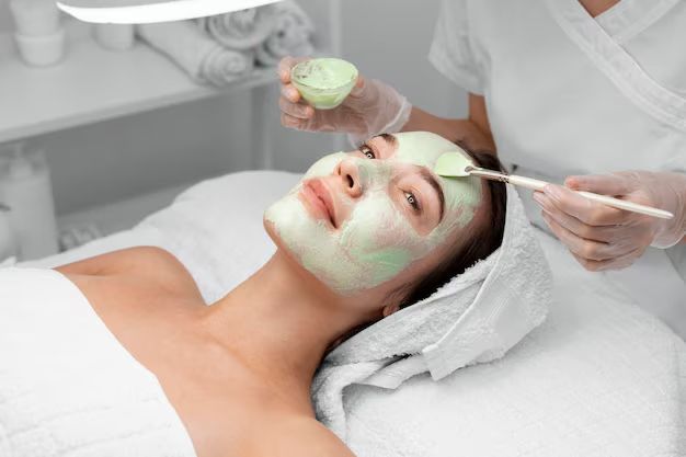 Woman receiving a green facial mask treatment at a beauty spa with therapist applying skincare product to face